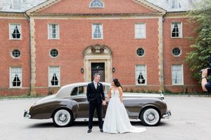 Classic brick manor house serves as elegant backdrop for newlyweds standing beside vintage Rolls Royce on wedding day.