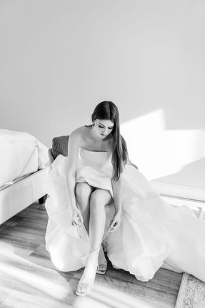 A bride in a white dress sits elegantly on a bed while adjusting her footwear in a sunlit room.