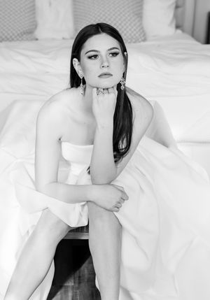 Black and white portrait of a subject in a white dress sitting on a bed with elegant pose and statement earrings.