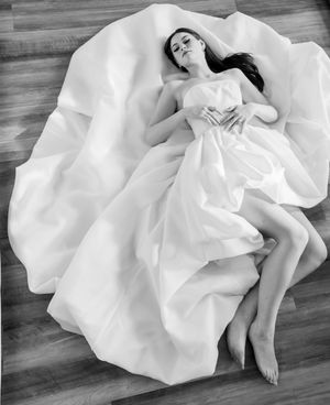 A figure in a flowing white dress lies gracefully on a wooden floor in an artistic black and white photograph.