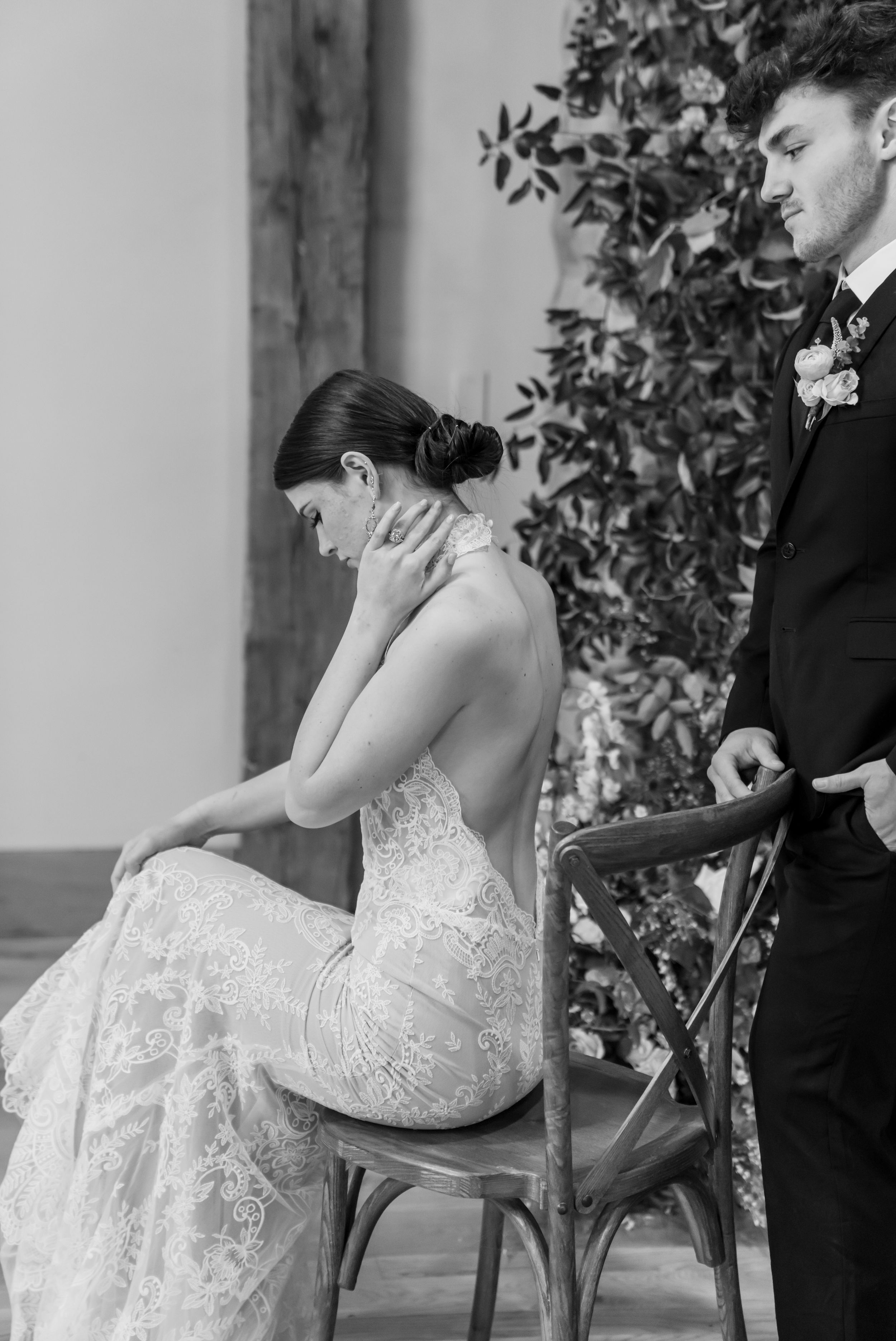 A touching black and white wedding moment with a bride in a lace dress seated on a wooden chair.