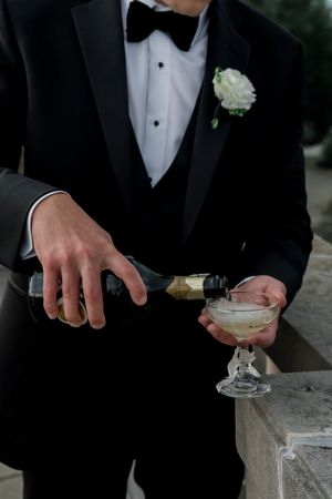 A groom in a black tuxedo and bowtie pours champagne into a coupe glass while wearing a white boutonniere.