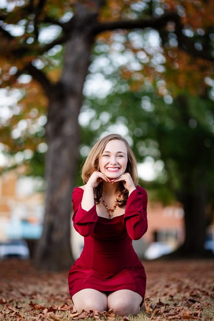 High school senior girl in a red dress kneeling in autumn leaves during a senior session in Harrison, Arkansas