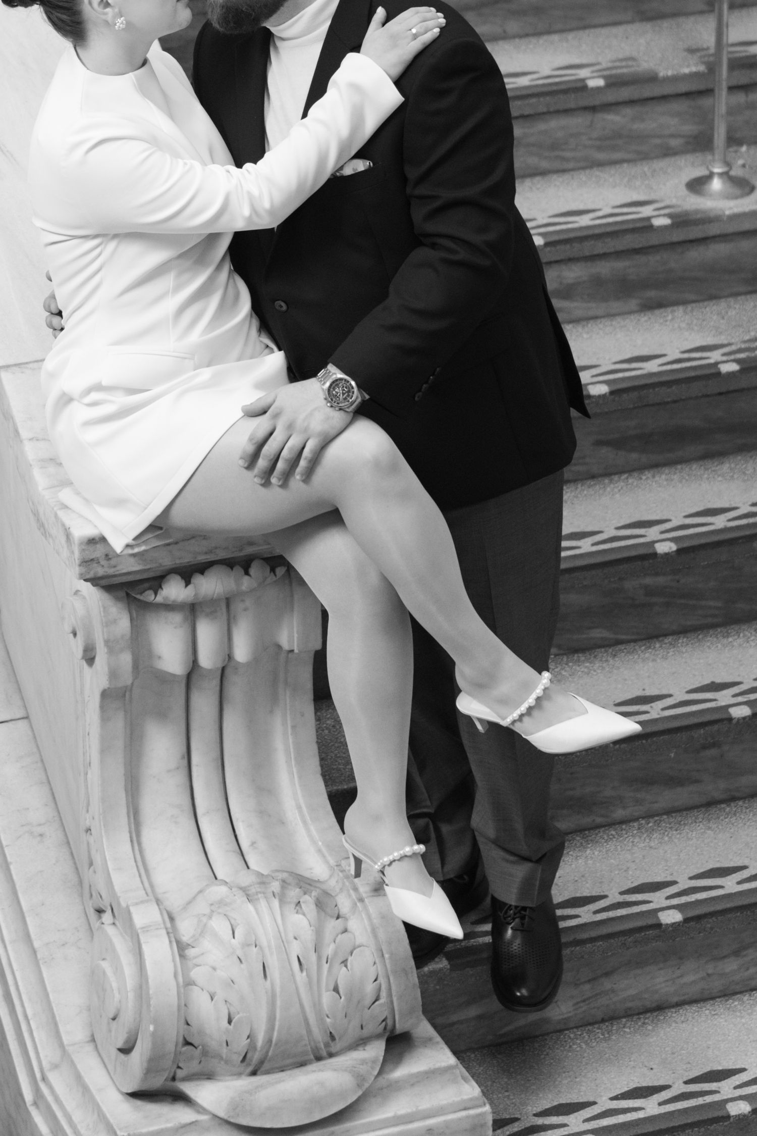 Black and white wedding photo of couple sitting on marble staircase balustrade, bride in white dress and heels.