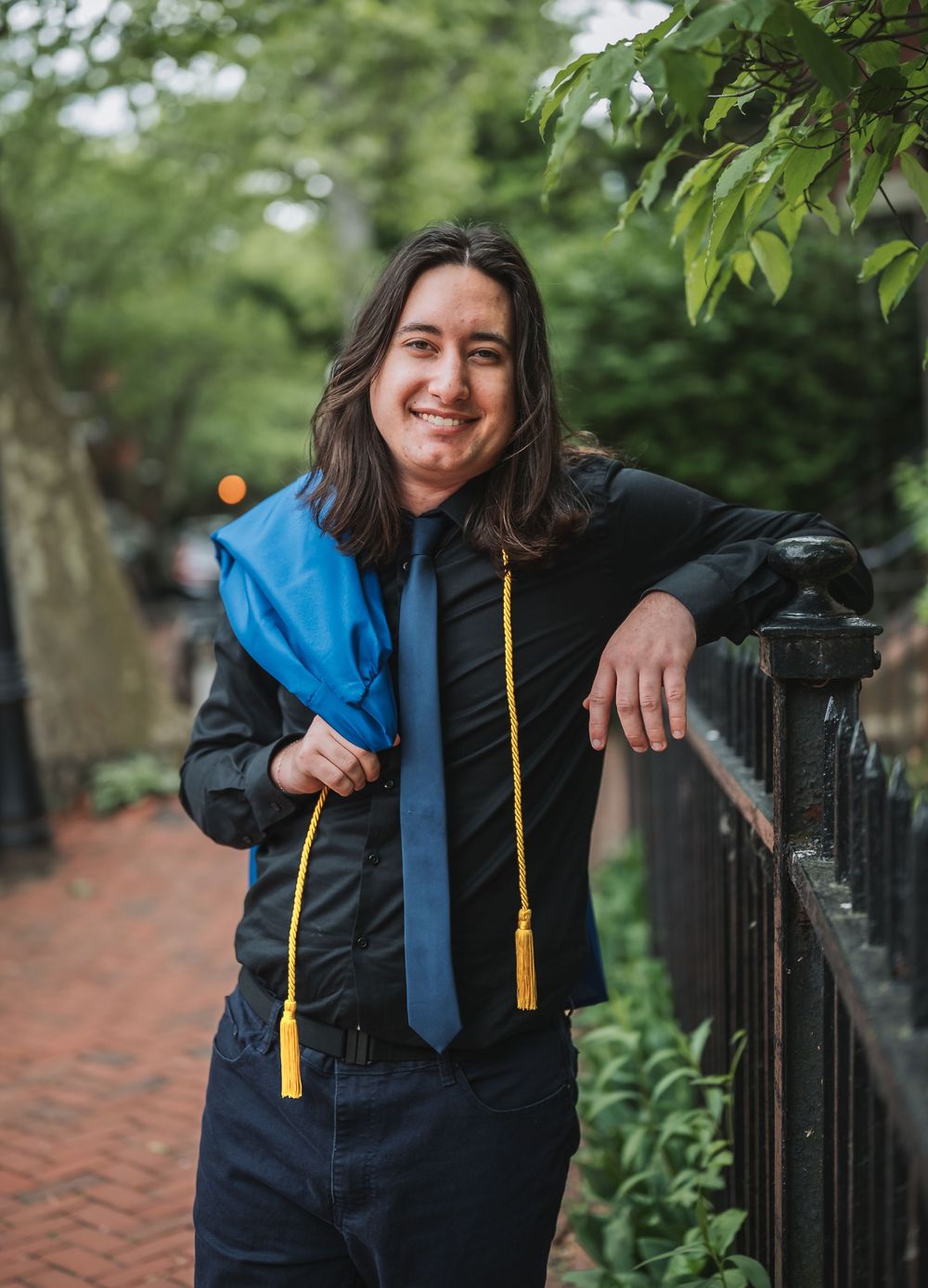 Smiling graduate poses outdoors holding blue cap with gold tassel, wearing black shirt and tie near iron fence.