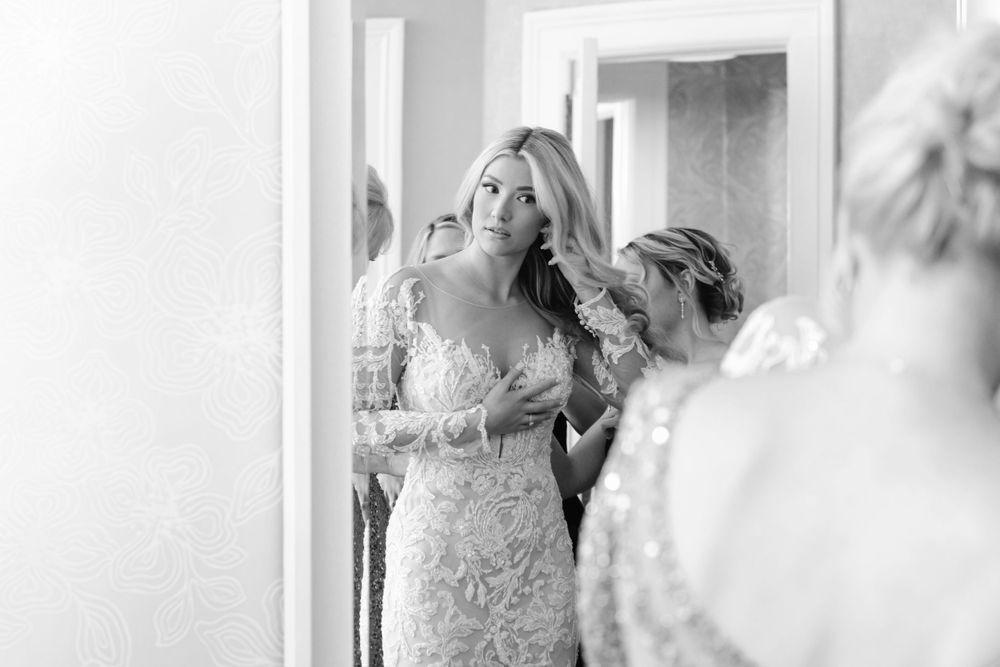A candid black and white wedding photo showing bride getting ready in front of a mirror.