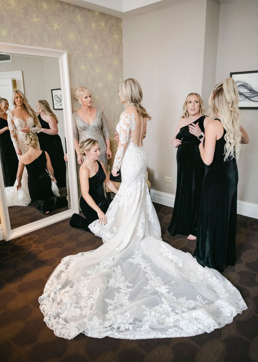 A bride in an elegant lace wedding gown with long train stands with bridesmaids in black dresses beside a large mirror.