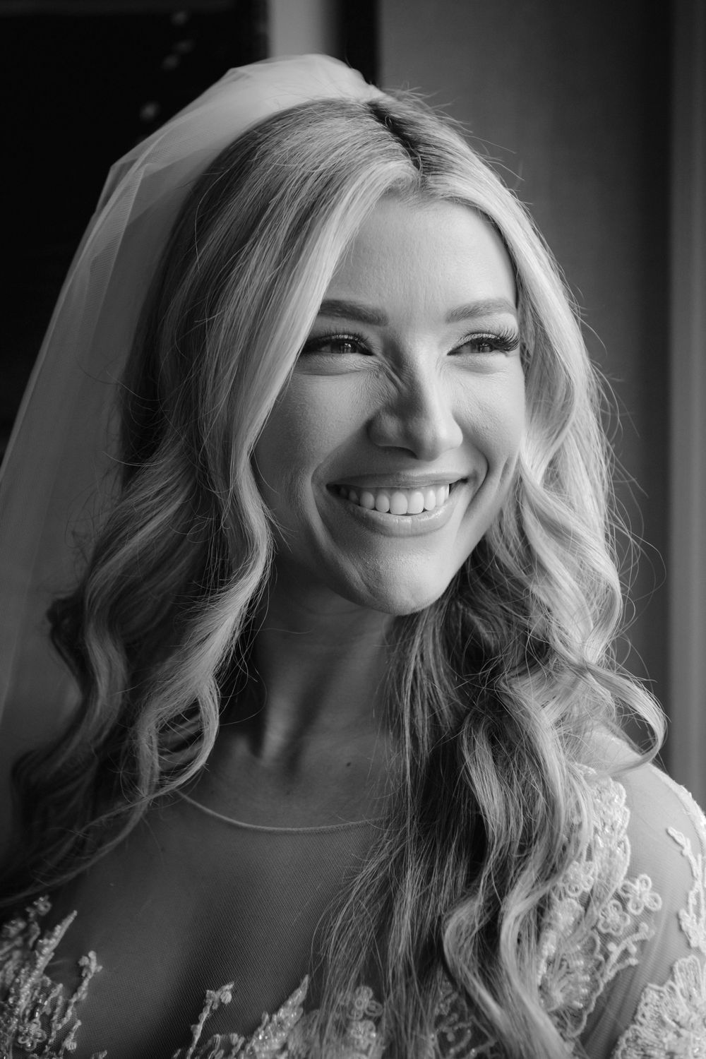 Black and white portrait of a radiant bride with flowing blonde waves wearing a lace wedding dress and veil showing a joyful smile.