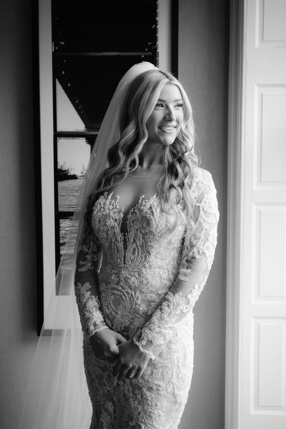 Black and white portrait of a bride in an elegant lace long-sleeve wedding gown with a veil, smiling by a window.