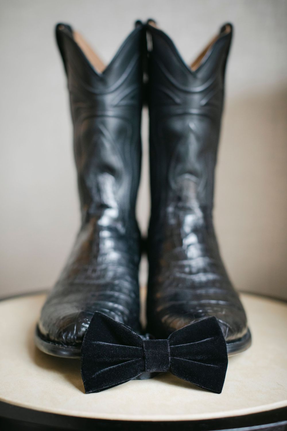 Black leather cowboy boots with a black velvet bow tie placed in front on a light surface.