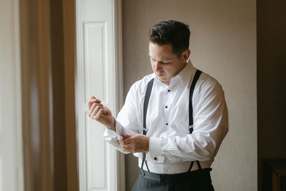 A groom in a classic white dress shirt and suspenders adjusts his cufflinks near a window.