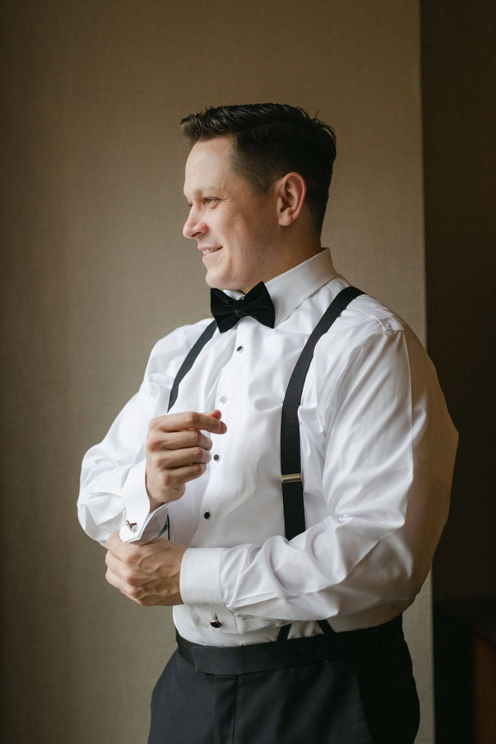 Well-dressed groom in white dress shirt and black suspenders adjusts cuff while looking to the side in elegant formal attire.