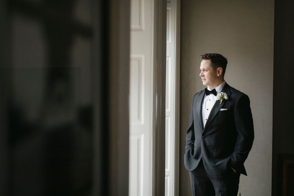 A groom in a classic black tuxedo stands pensively by a window in natural light.