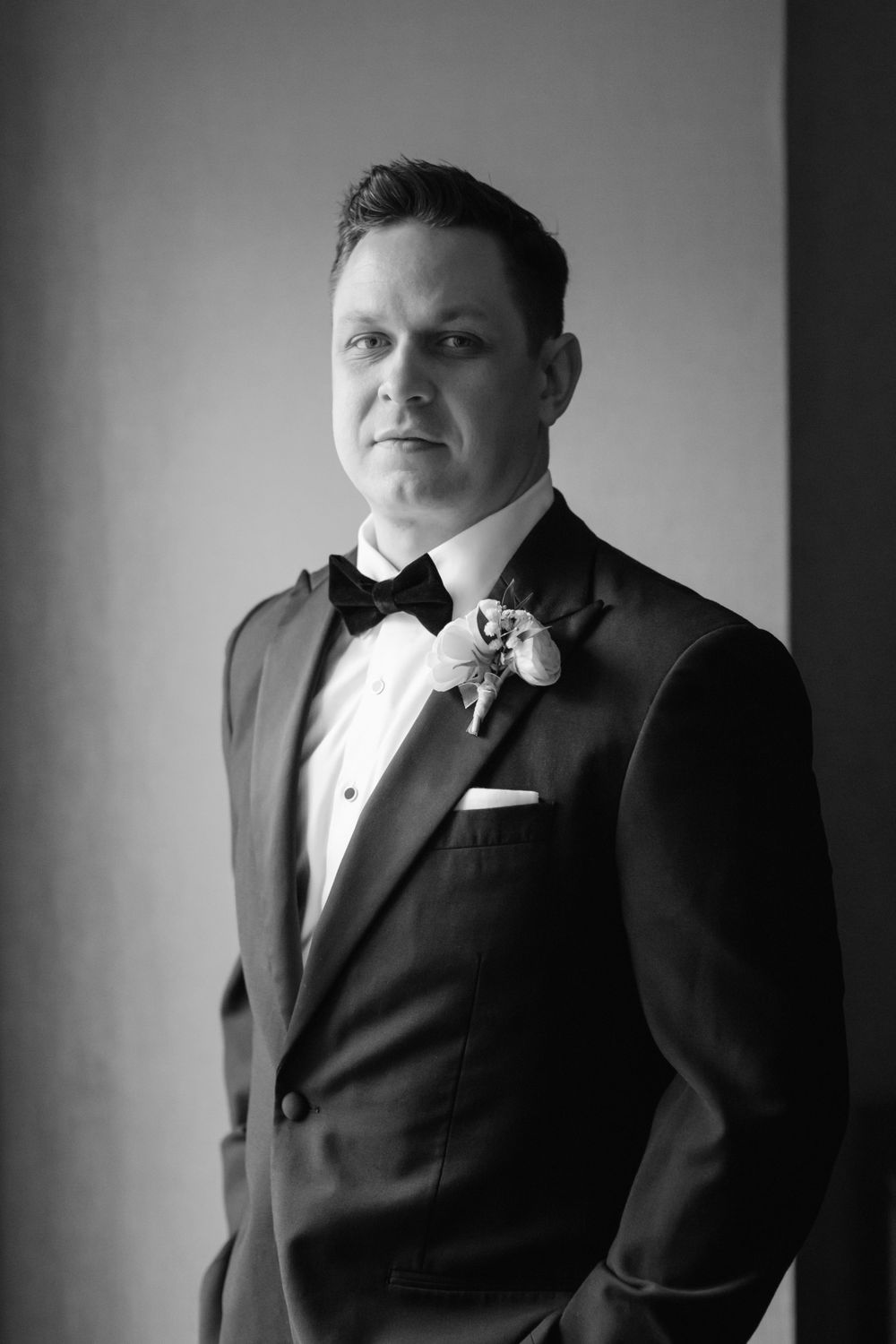 Black and white portrait of a groom in formal tuxedo with bow tie and boutonniere in elegant lighting.