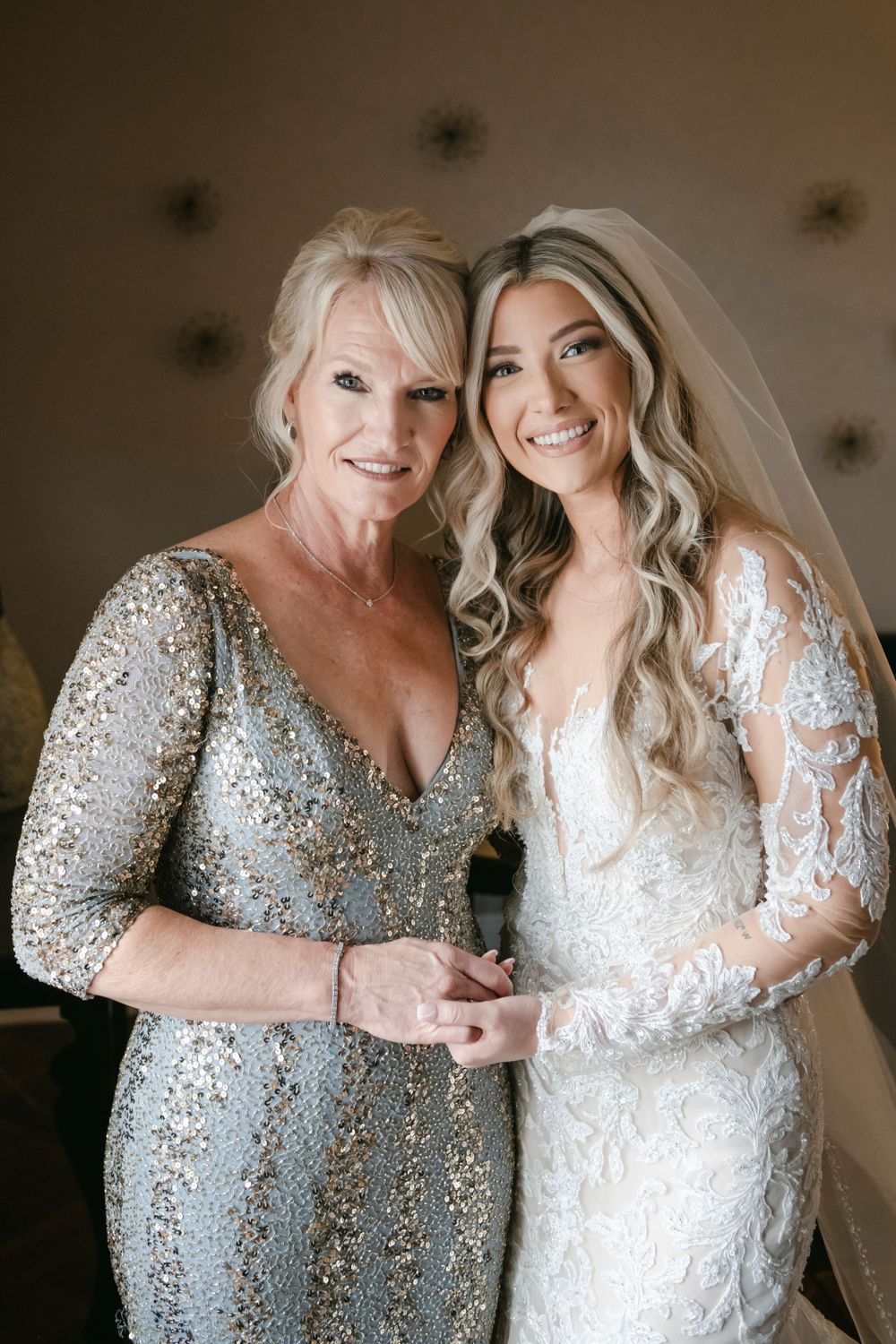 A special moment between a bride and her mother wearing sparkling formal gowns in silver and white lace on a wedding day.