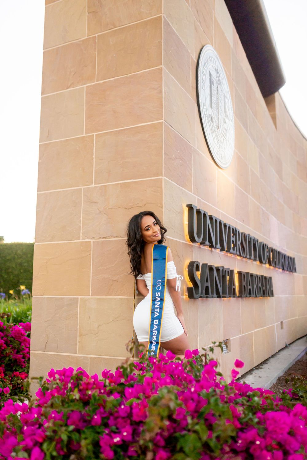 Vibrant pink flowers bloom beneath a university building sign with stone walls.