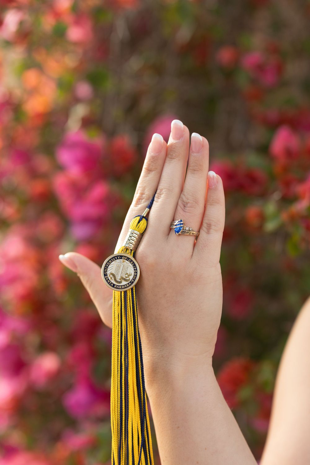 A hand wearing a ring holds a yellow graduation tassel against a blurred pink floral background.