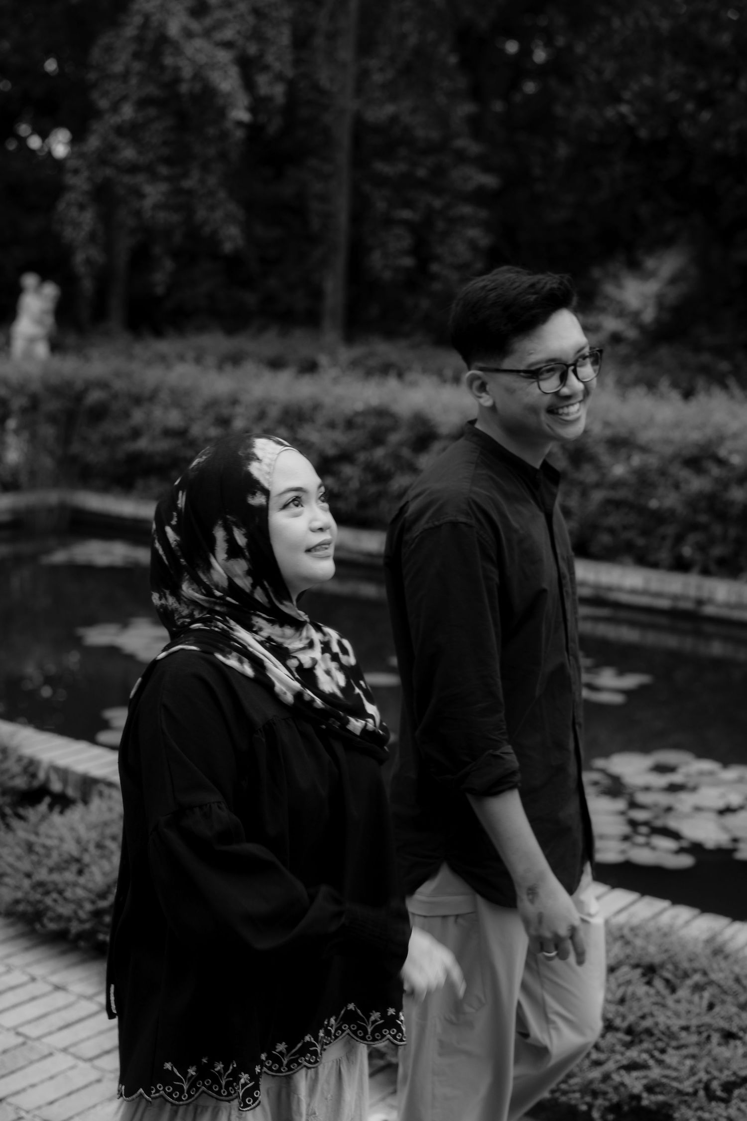Black and white photo of a smiling couple holding hands in a garden, woman wearing hijab and man in glasses.
