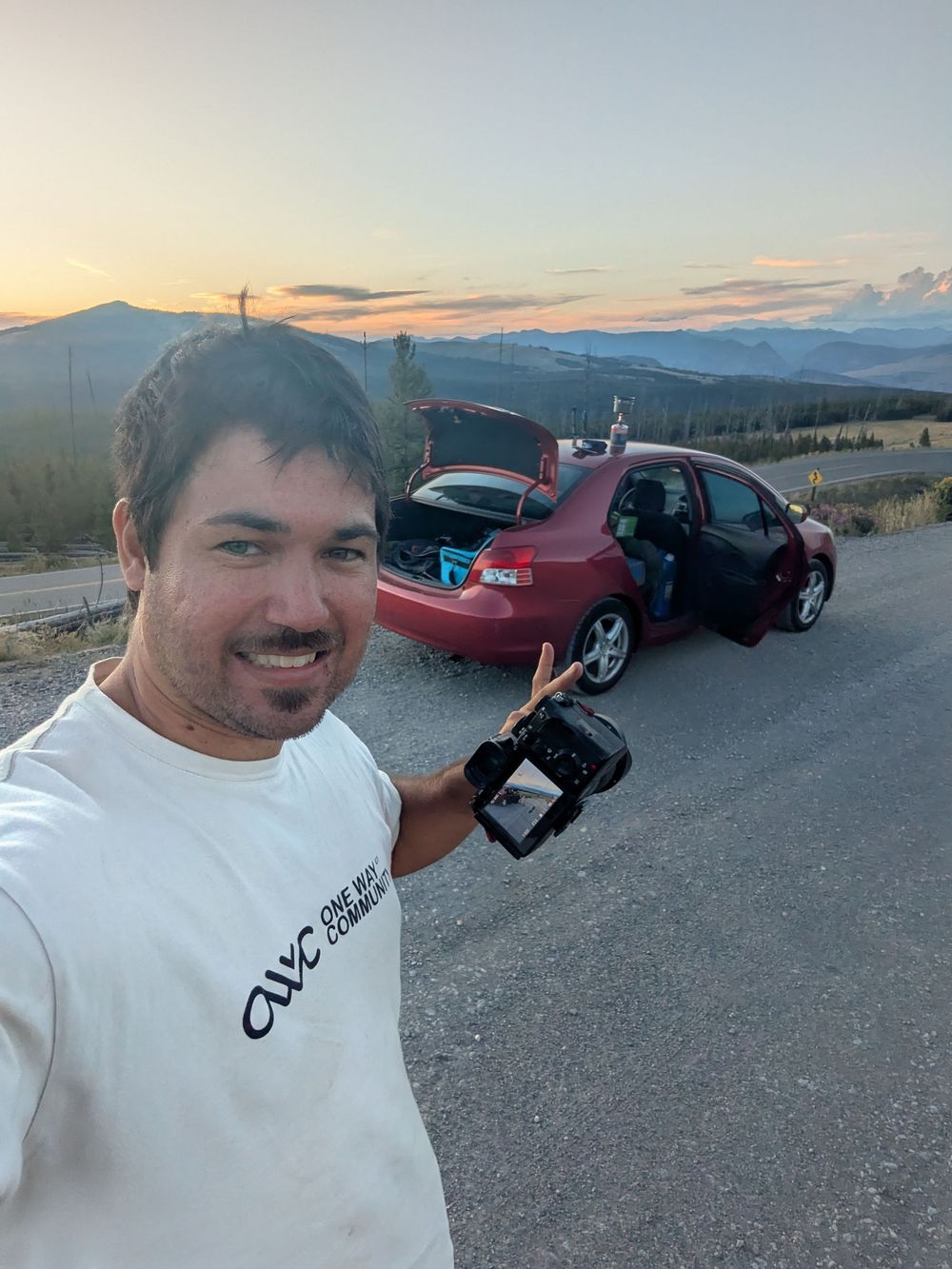 A series of sports cars parked along a scenic mountain road at sunset with distant peaks in view.