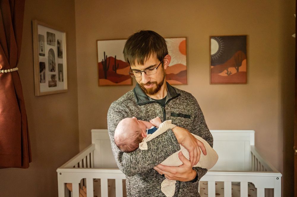 Parent in a gray sweater holds and comforts a newborn baby in a warmly lit room with framed artwork on the walls.