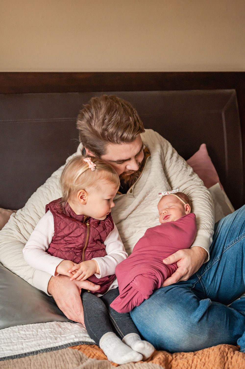 A parent in a cream sweater cuddles with a toddler in burgundy and a newborn in pink on a cozy couch.