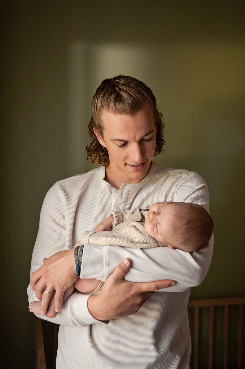 A person in a white sweater tenderly holds and gazes at a sleeping newborn baby wrapped in a light-colored blanket.