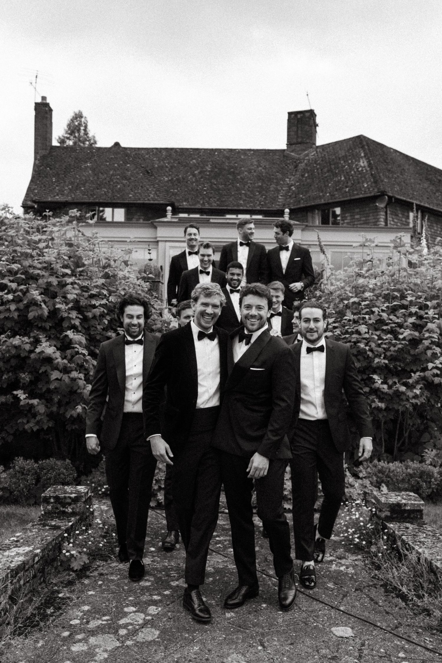 A group of men in formal black tuxedos pose together in a garden outside a rustic house.