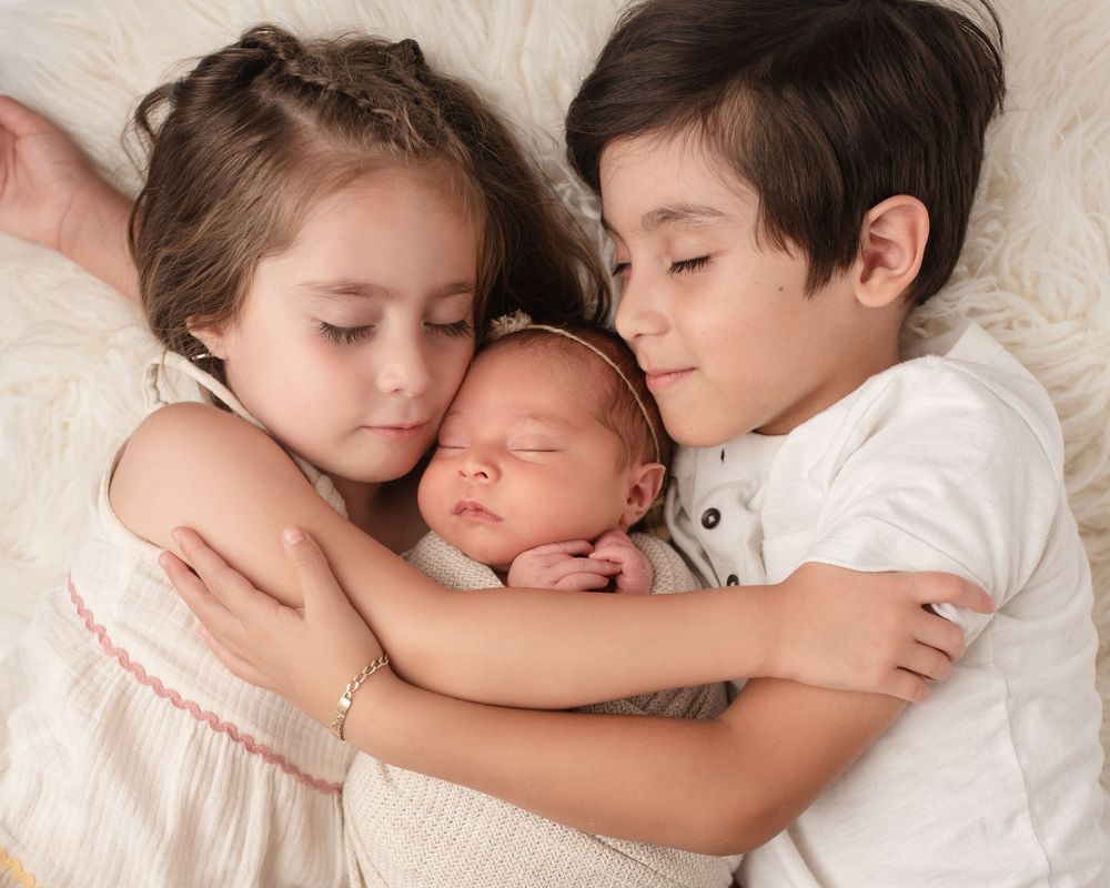 siblings cuddling newborn on white flokati rug.