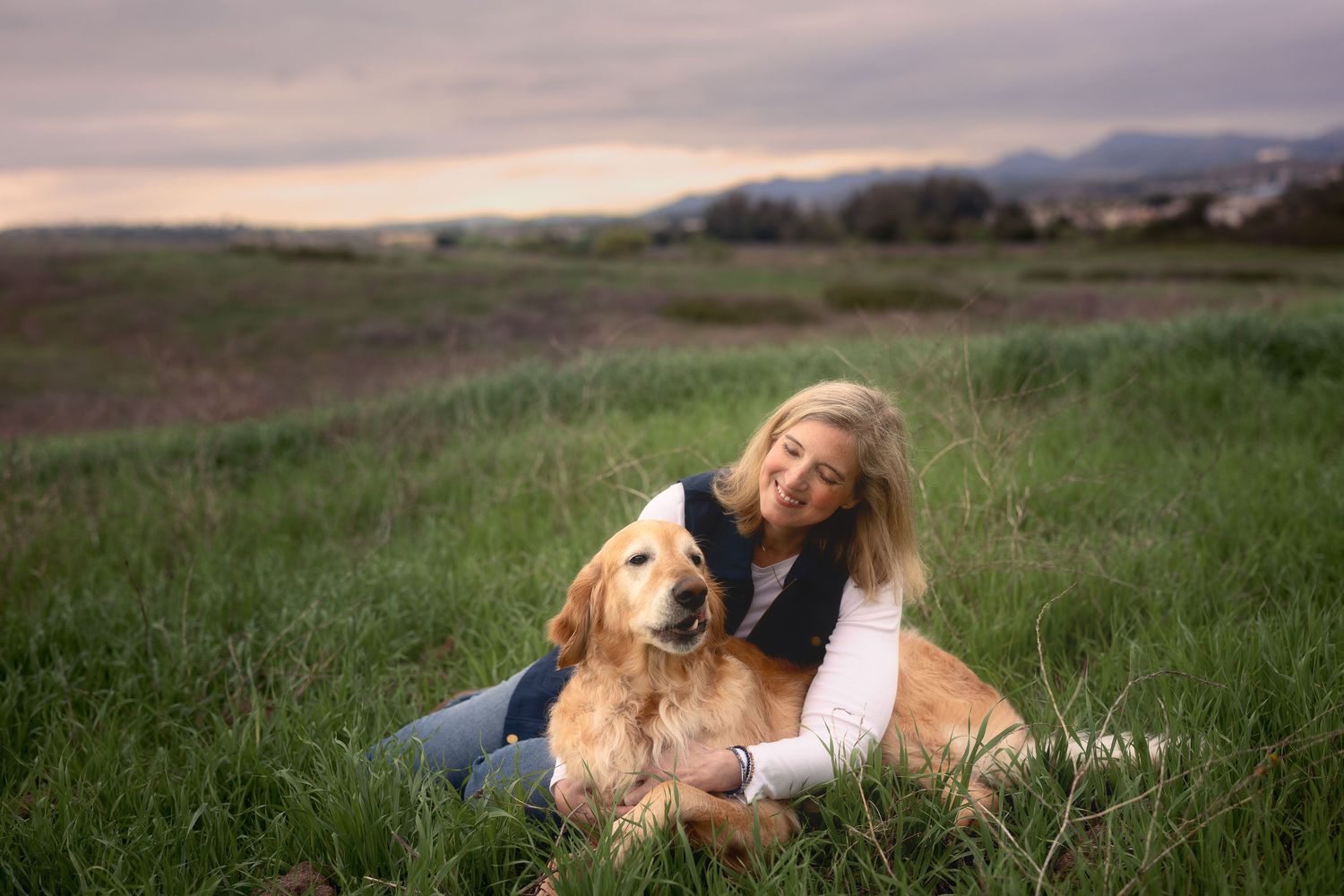 A person and Golden Retriever dog relax together in a grassy field at sunset with mountains in the background.