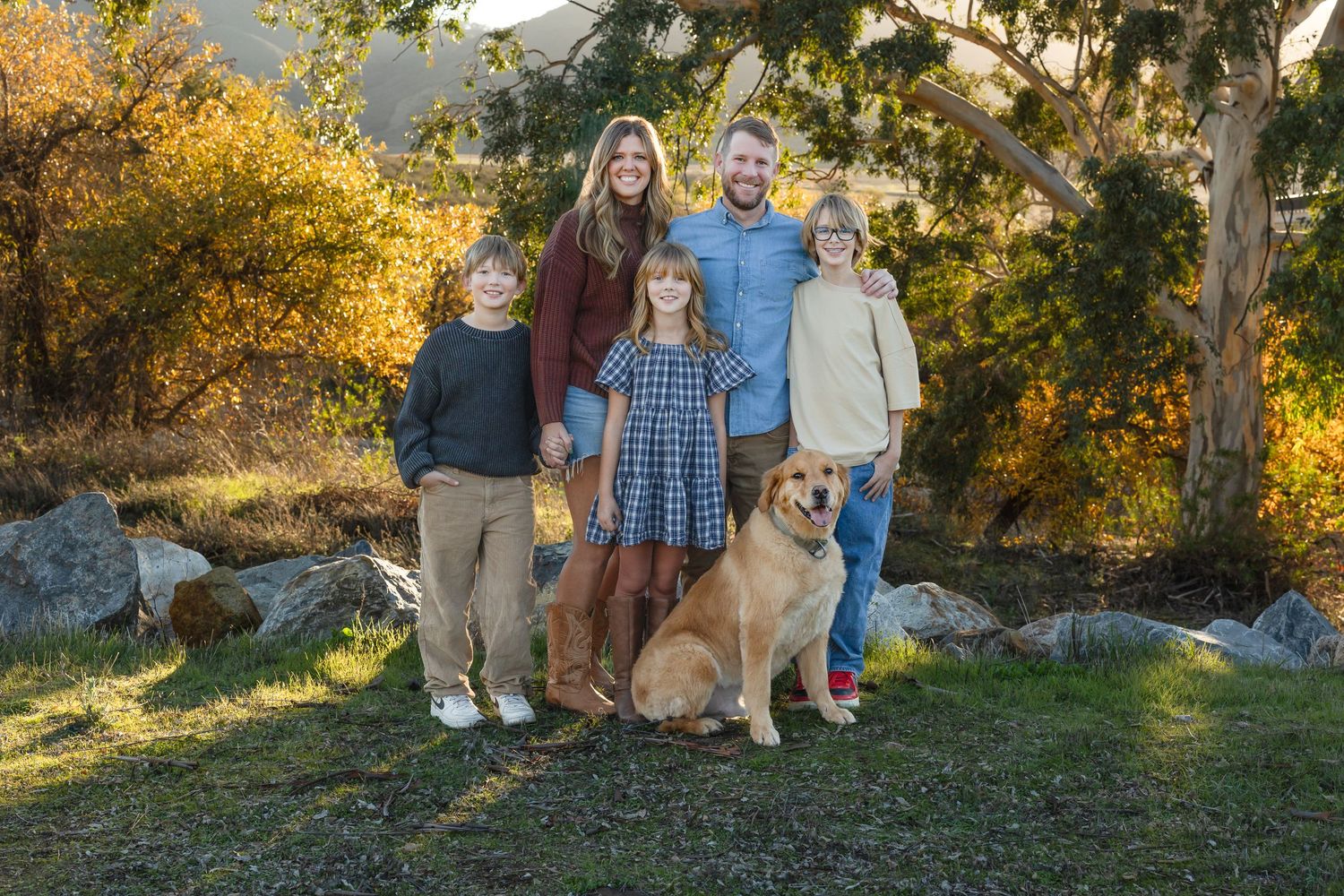 Family and golden retriever dog pose together in autumn outdoor setting with golden foliage in background.