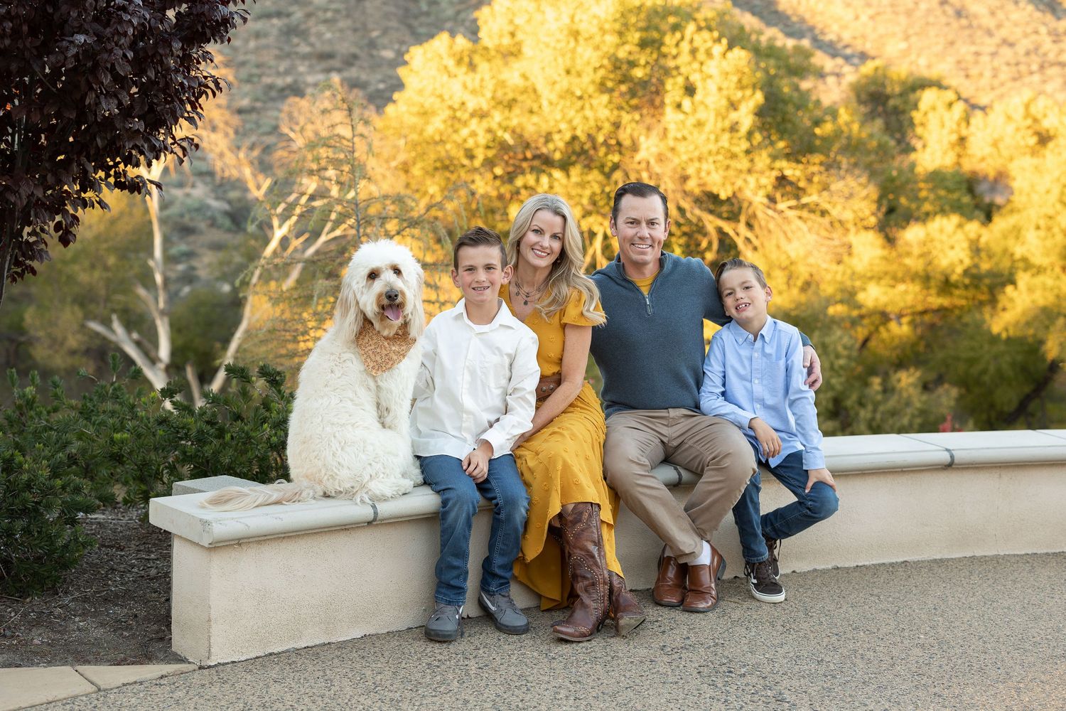 Group sitting on stone wall with golden autumn foliage and trees in background during sunset.
