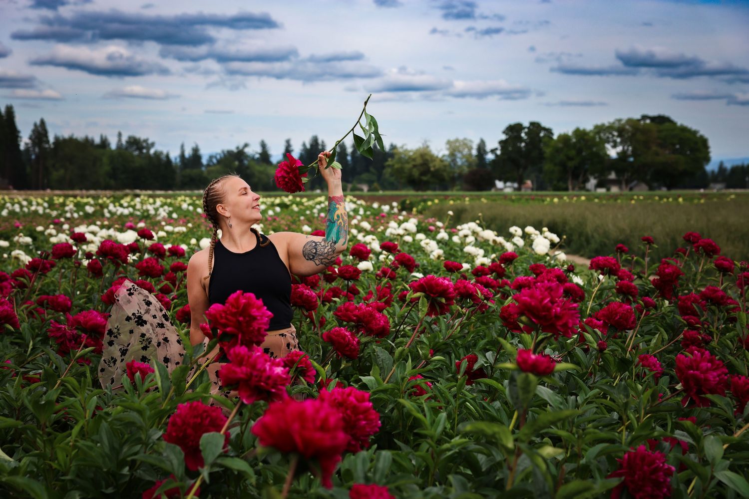 Brigit, photographer, in a black tank top amid vibrant red and white peony flowers in a blooming garden field.