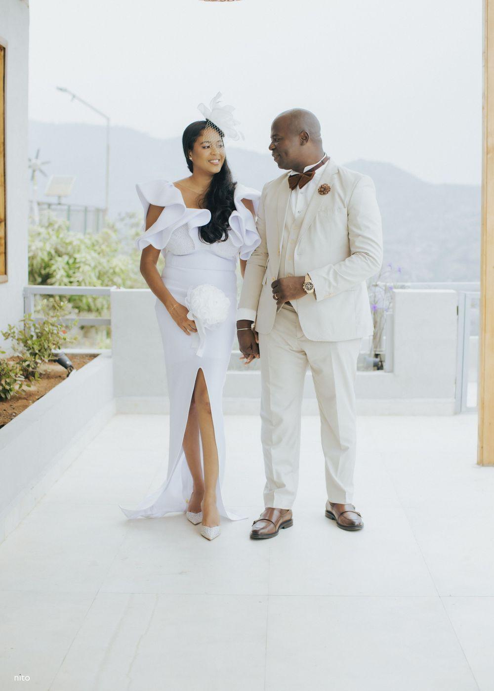 A stylish couple in white wedding attire poses playfully on a bright outdoor patio.