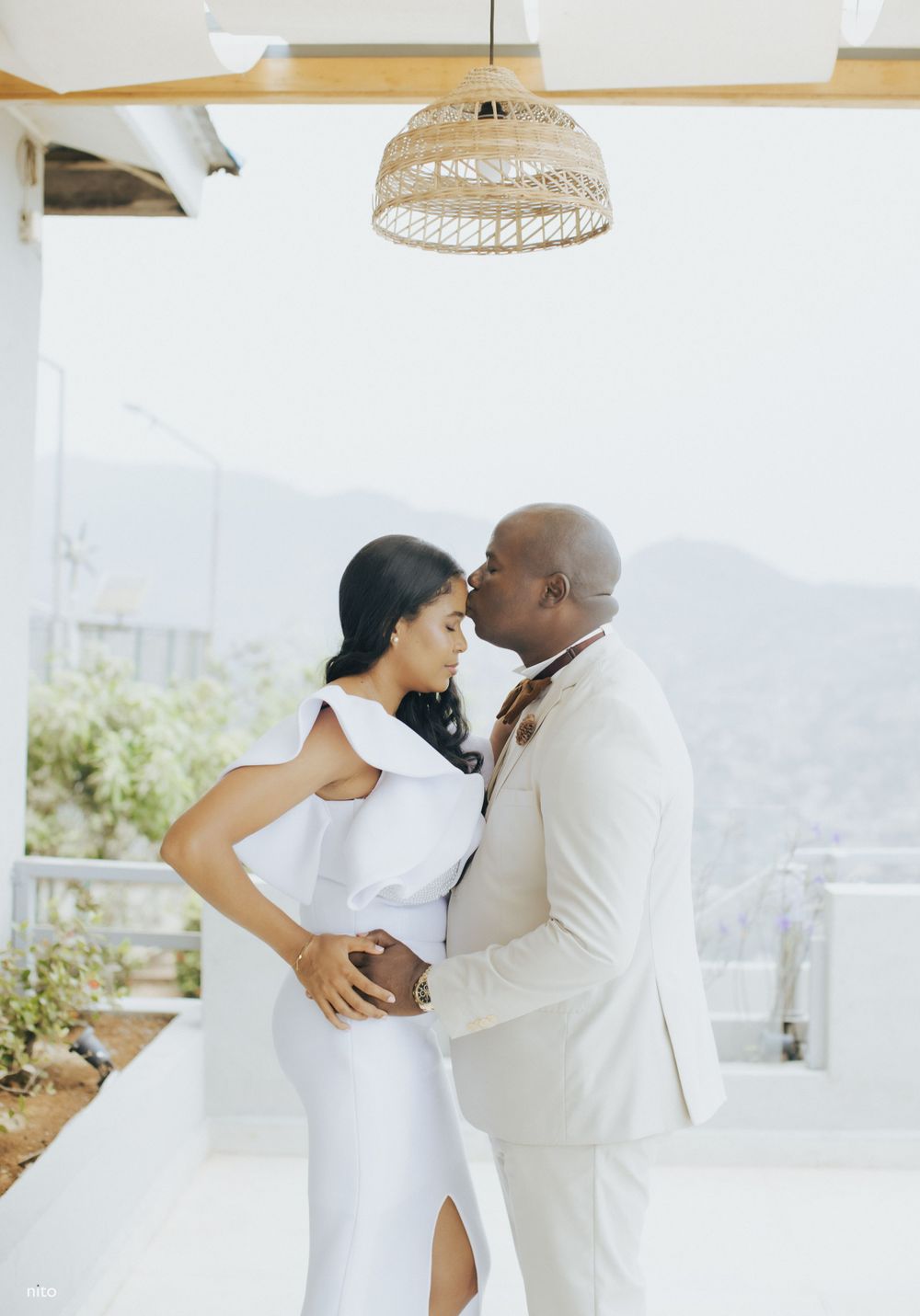Newlyweds in elegant white attire embrace under a woven pendant light with mountain views.