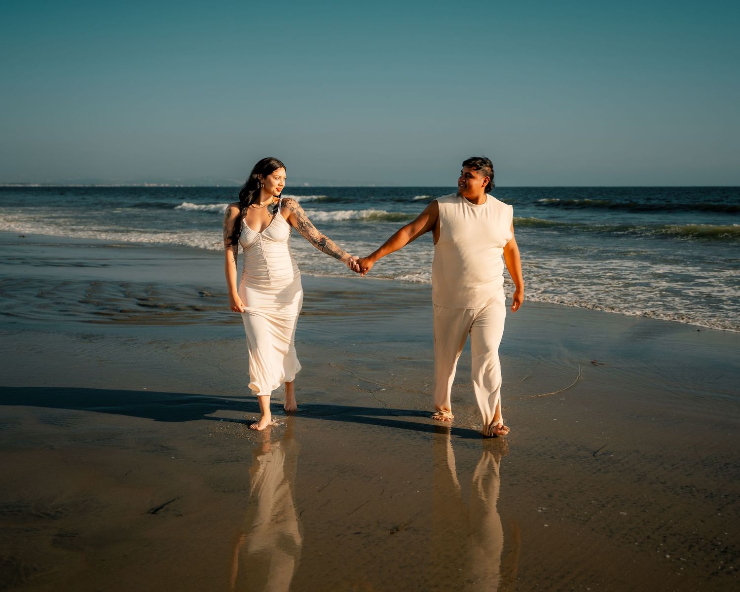 Romantic beach photoshoot of couple in white outfits walking along the shoreline at sunset.