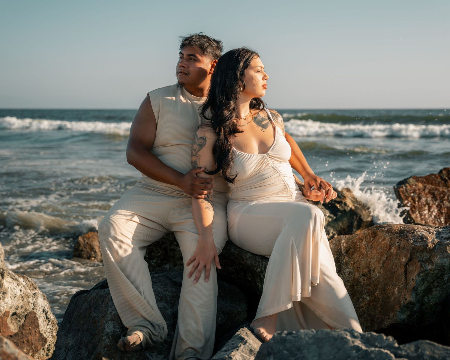 Couple in white clothing posing on rocky beach with ocean waves in background.