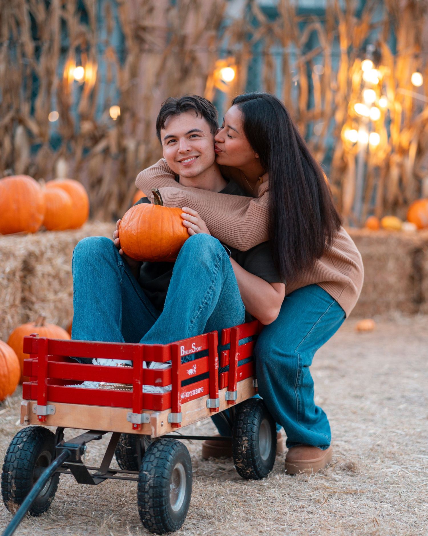 Romantic couple sharing intimate moment on red wagon surrounded by fall pumpkins.