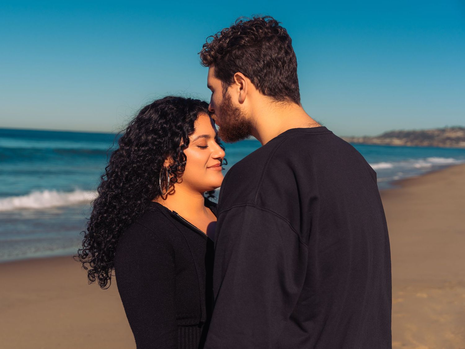 Romantic couple embraces on sunny beach with waves and palm trees in background.