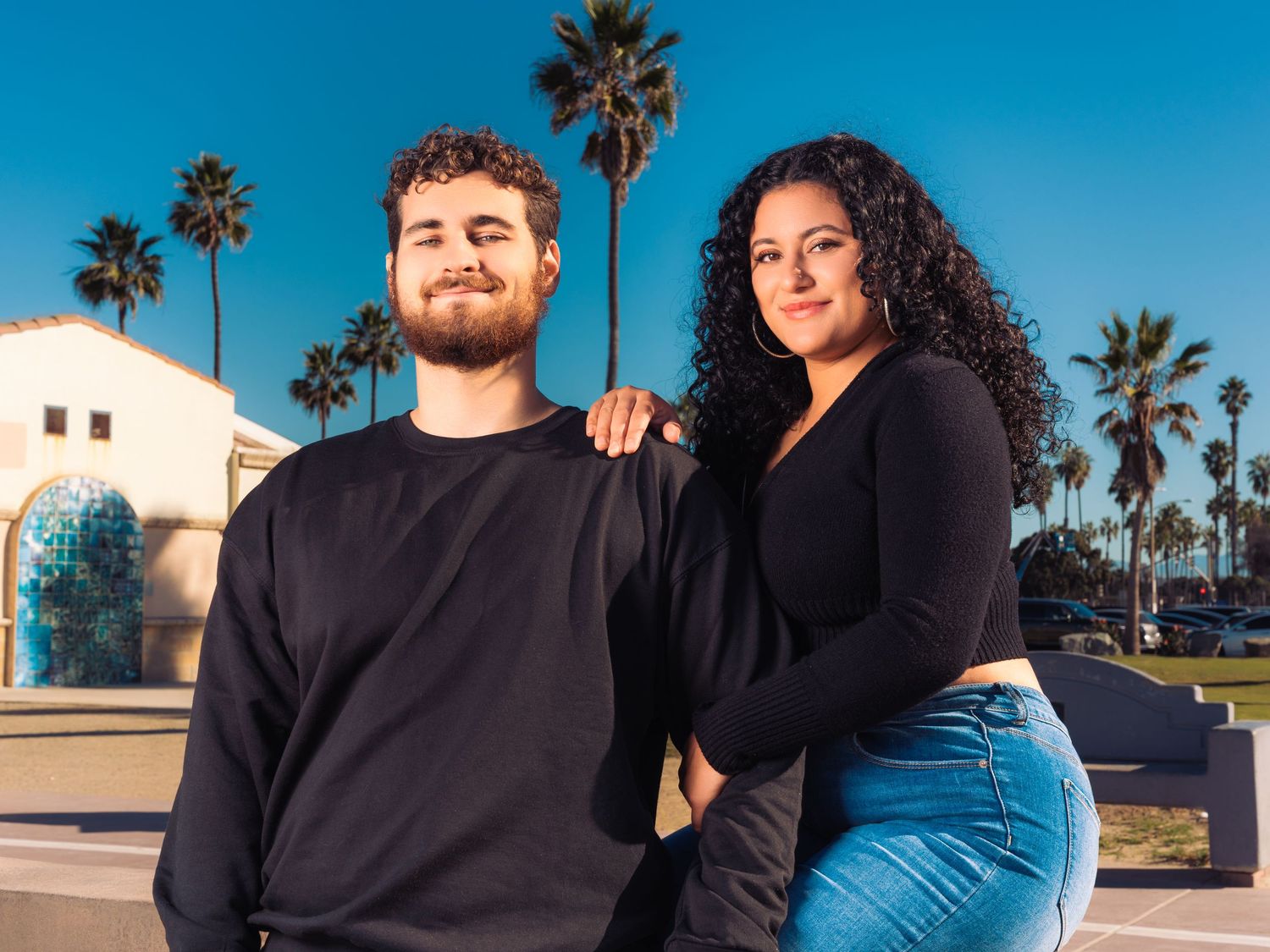 Couple in black clothing shares intimate moment by palm trees on California beach.