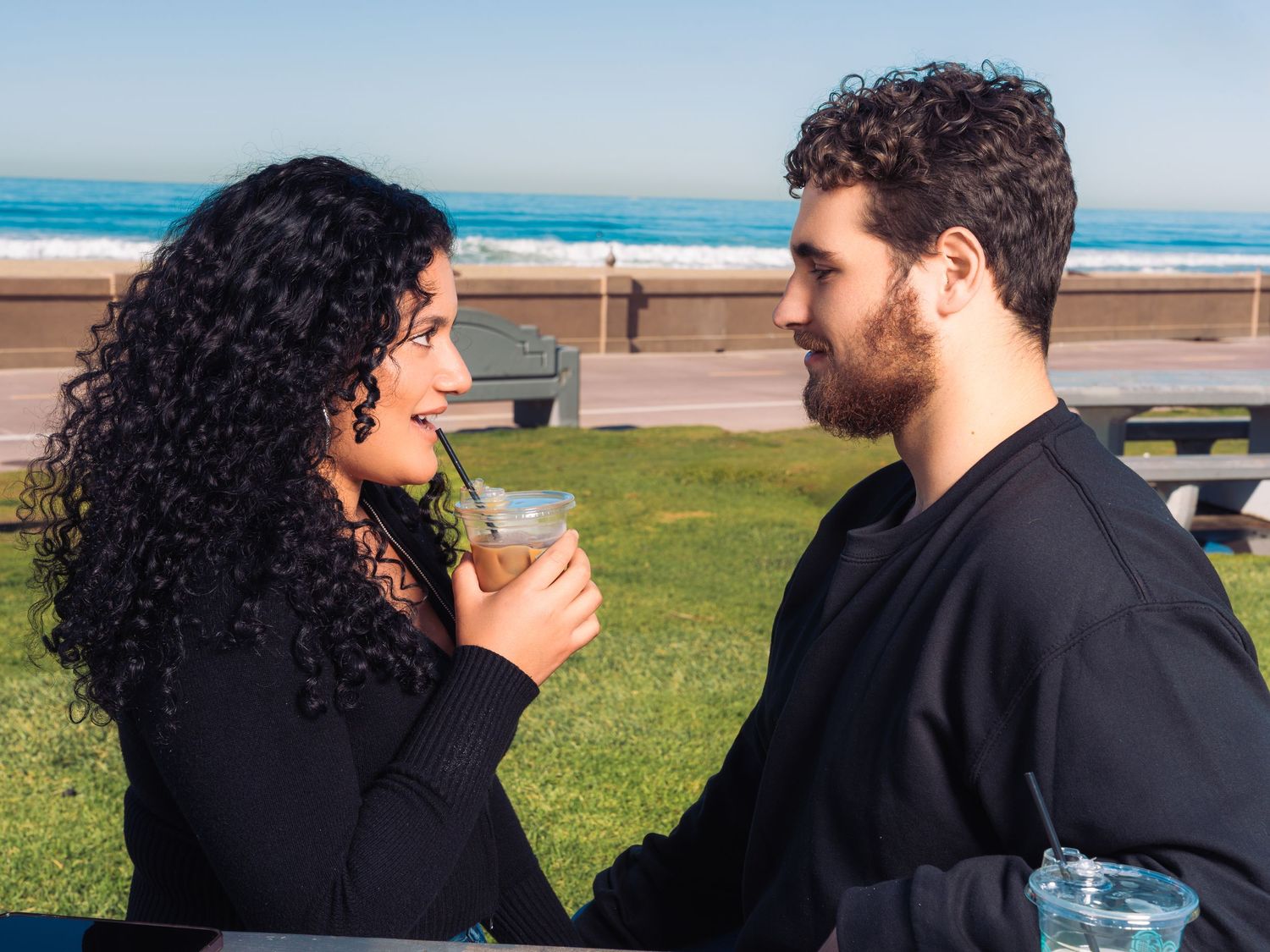 Couple in dark clothing embraces near grassy oceanfront area with blue skies.