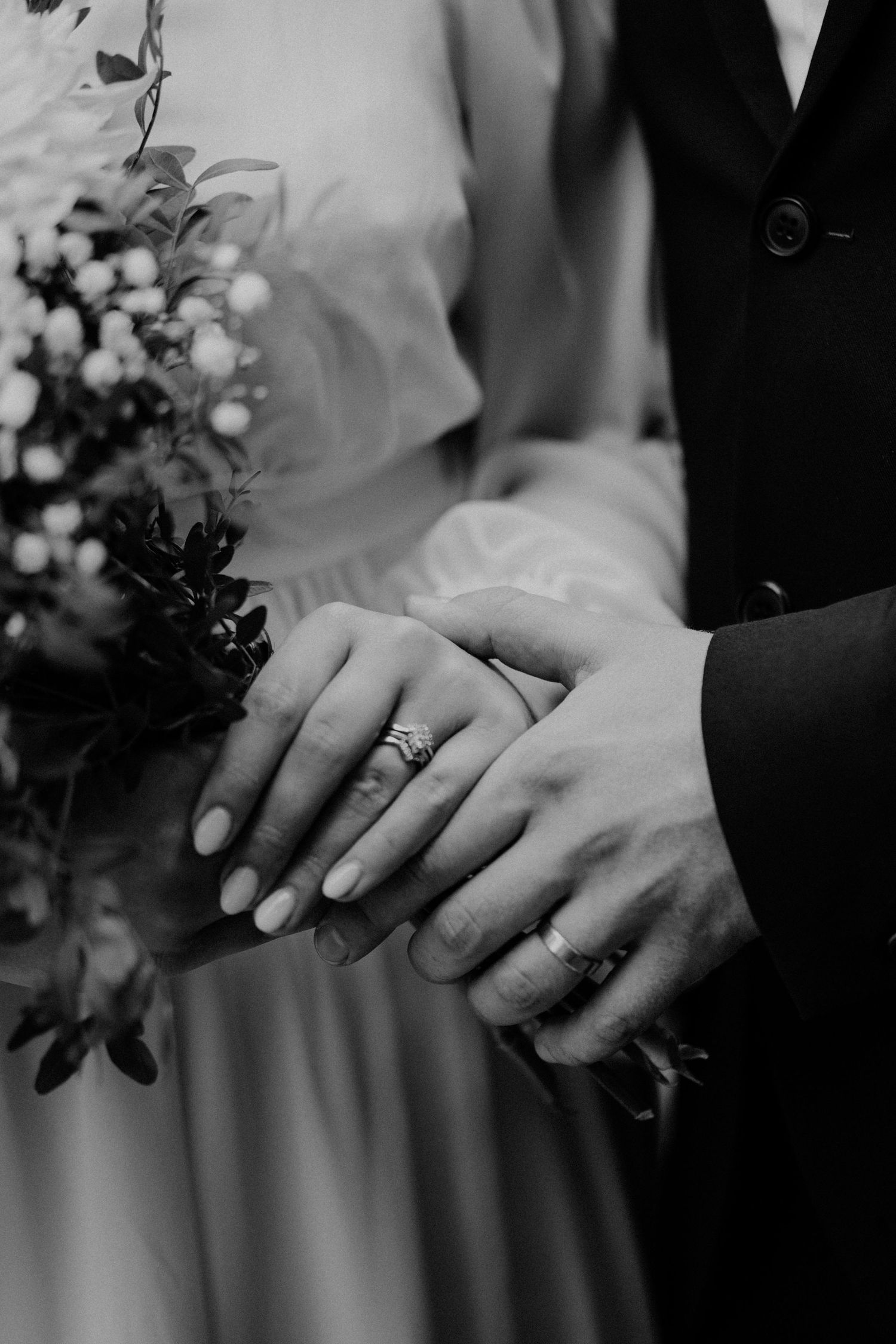 Black and white close-up of wedding rings and hands holding bridal bouquet.