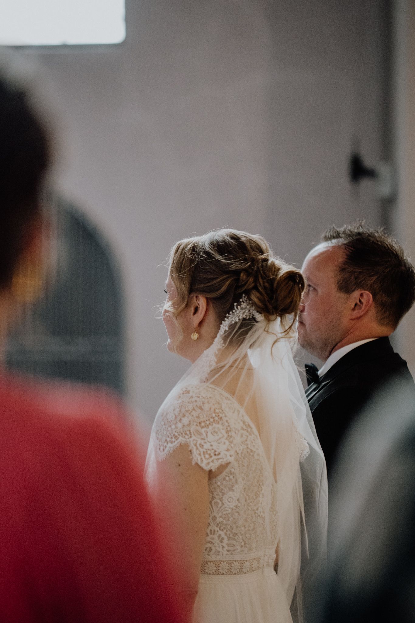 Side profile of a couple during their wedding ceremony, bride wearing a lace dress with veil and groom in black suit.