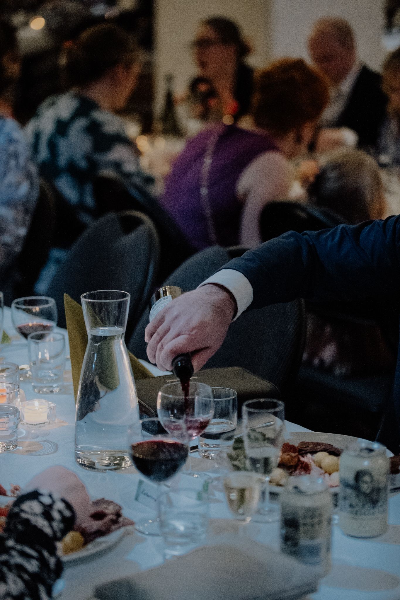 Candlelit dinner table with wine glasses and people's hands reaching for food and drinks in dim lighting.