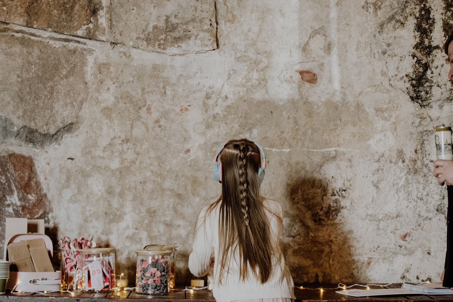 Person with long brown hair sits at rustic desk with vintage wall backdrop and scattered books and papers.