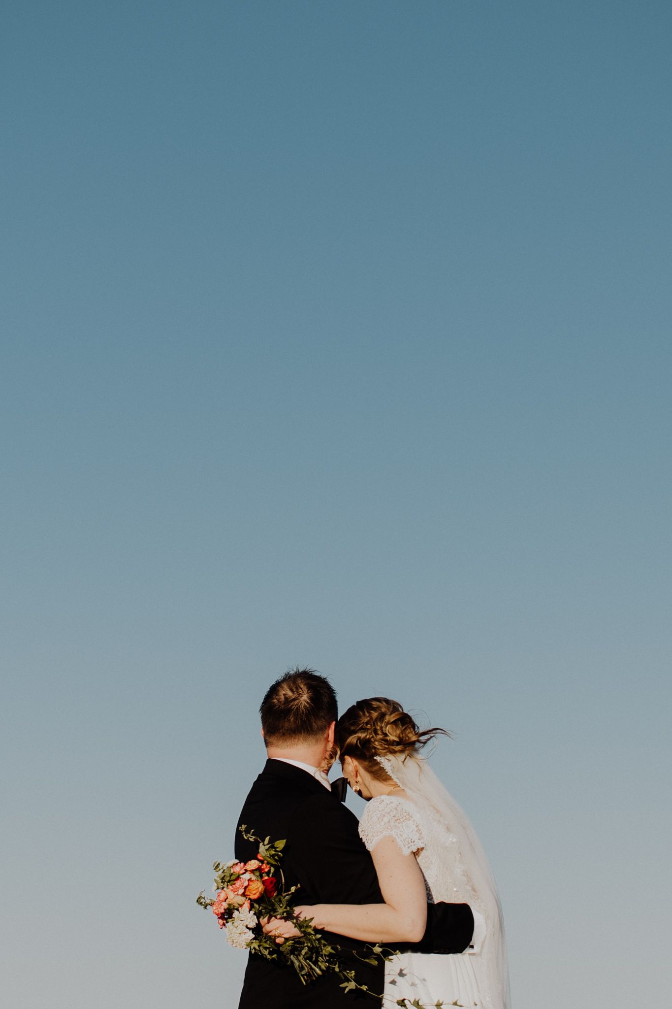A couple in wedding attire embraces during a romantic sunset moment against a blue sky backdrop.