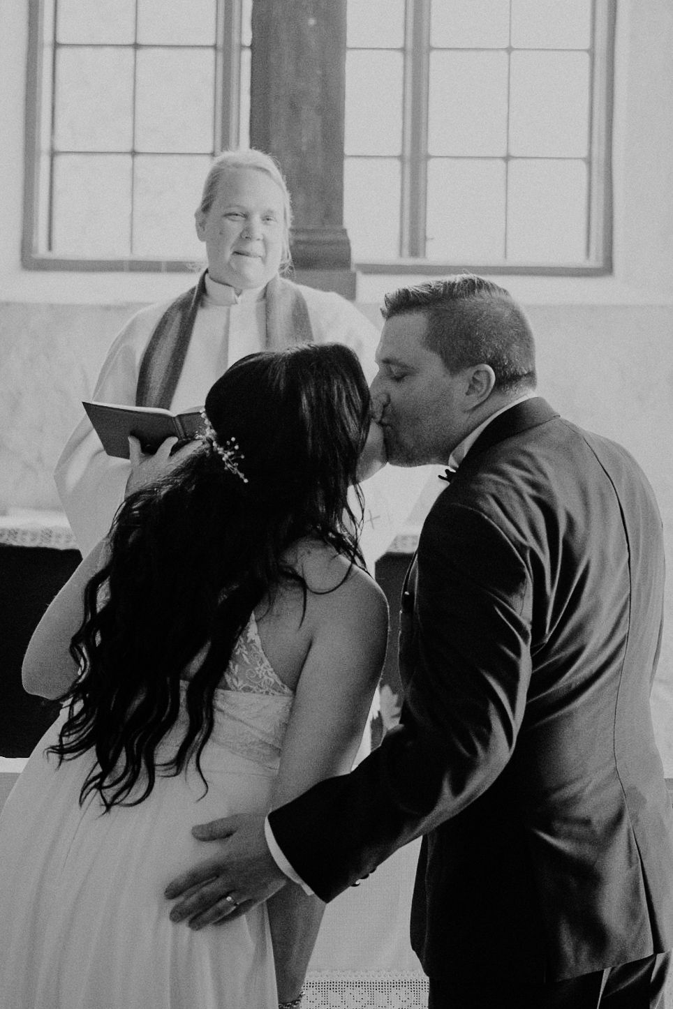 A bride and groom share their first kiss at the altar during a black and white church wedding ceremony.