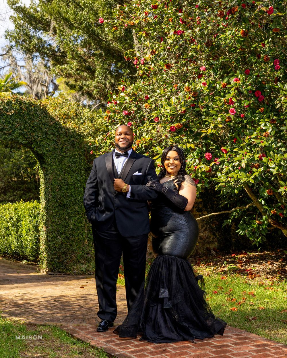 A couple poses together in formal attire among blooming red camellias and manicured garden paths on a sunny day.