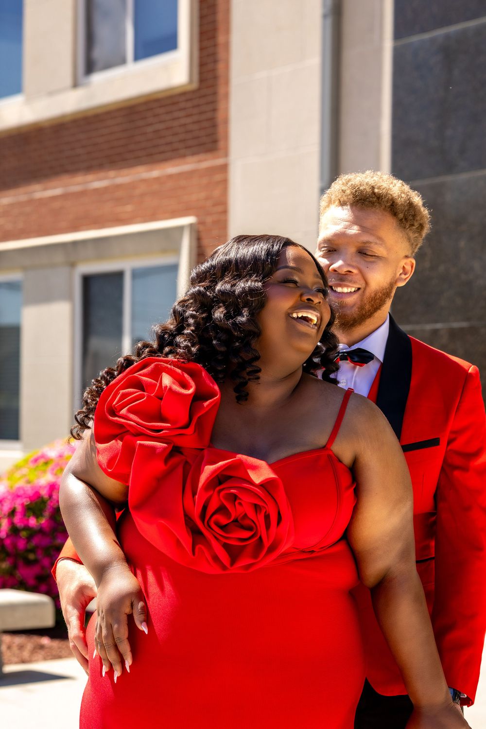 A couple wearing matching red formal attire share a joyful moment together outdoors in the sunshine.