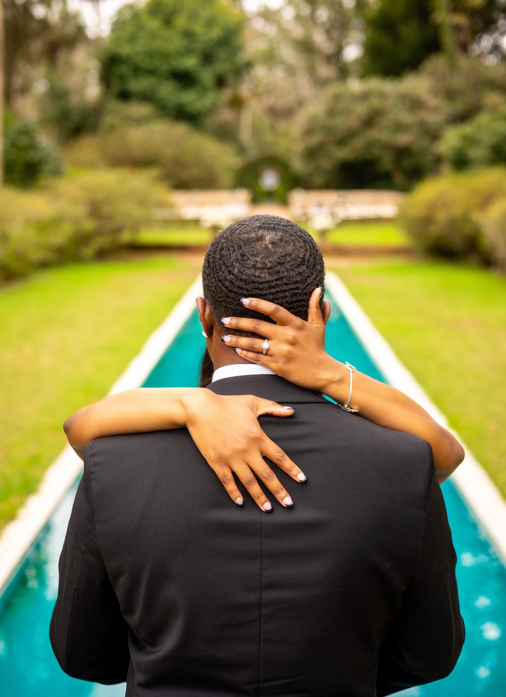 A couple embraces near a reflecting pool with lush green gardens in the background, shot from behind.
