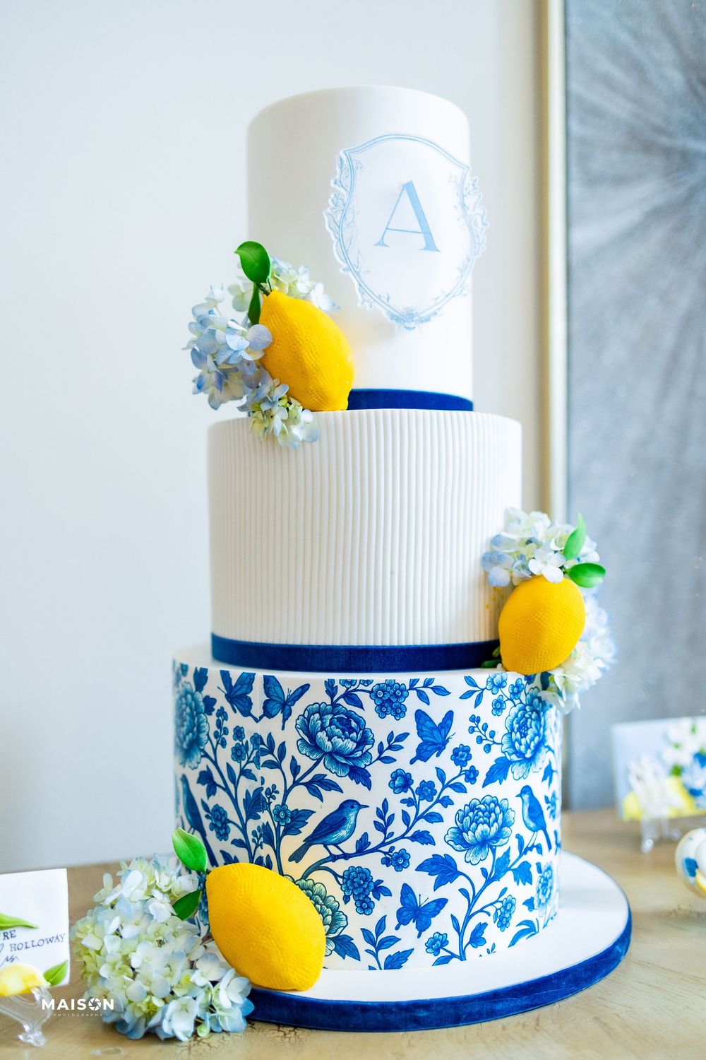 Elegant white wedding cake decorated with blue floral pattern, yellow lemons and white flowers on display.